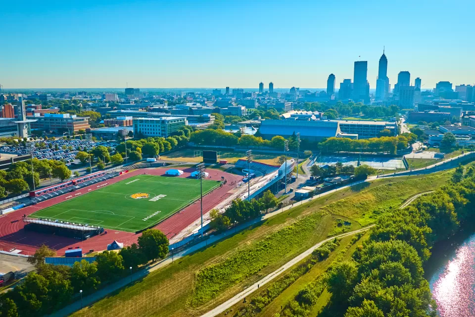 Aerial view of a bright green turf soccer stadium surrounded by red surfaced track with a river to the right and a downown skyline in the background against a bright blue sky.